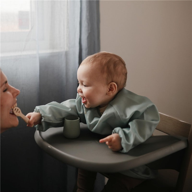 Mushie Long Sleeve Bib in Roman Green worn by a smiling baby reaching for a wooden spoon, seated at a high chair.