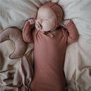 Infant peacefully sleeping in a blush-colored outfit and matching bonnet, with a pacifier and a crescent-shaped plush pillow nearby.