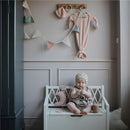 A baby sits on a white bench, holding a plush toy, surrounded by pastel decor, including a pink outfit and decorative bunting.