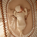 A sleeping baby in a cream-colored outfit and bonnet, nestled in a cozy cradle with a star-shaped pillow and a wooden teething toy.