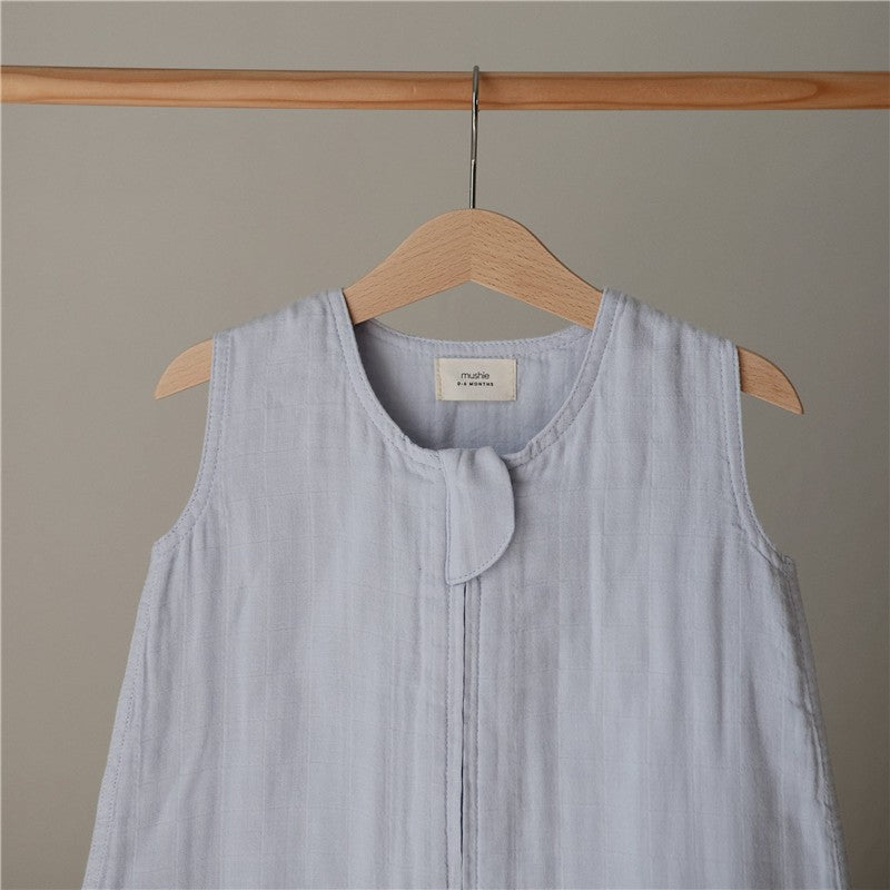 Mushie Muslin Sleep Bag in baby blue, featuring a front button closure and hanging on a wooden hanger.