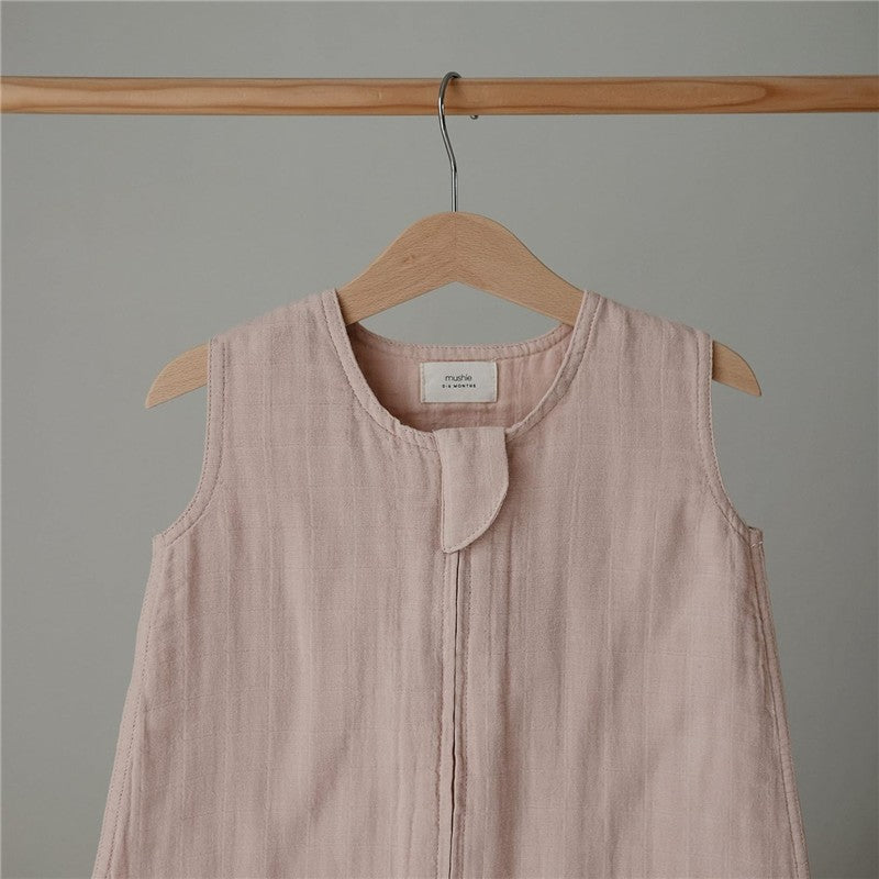 Mushie Muslin Sleep Bag in blush color, featuring a round neckline and front snap closure, displayed on a wooden hanger.