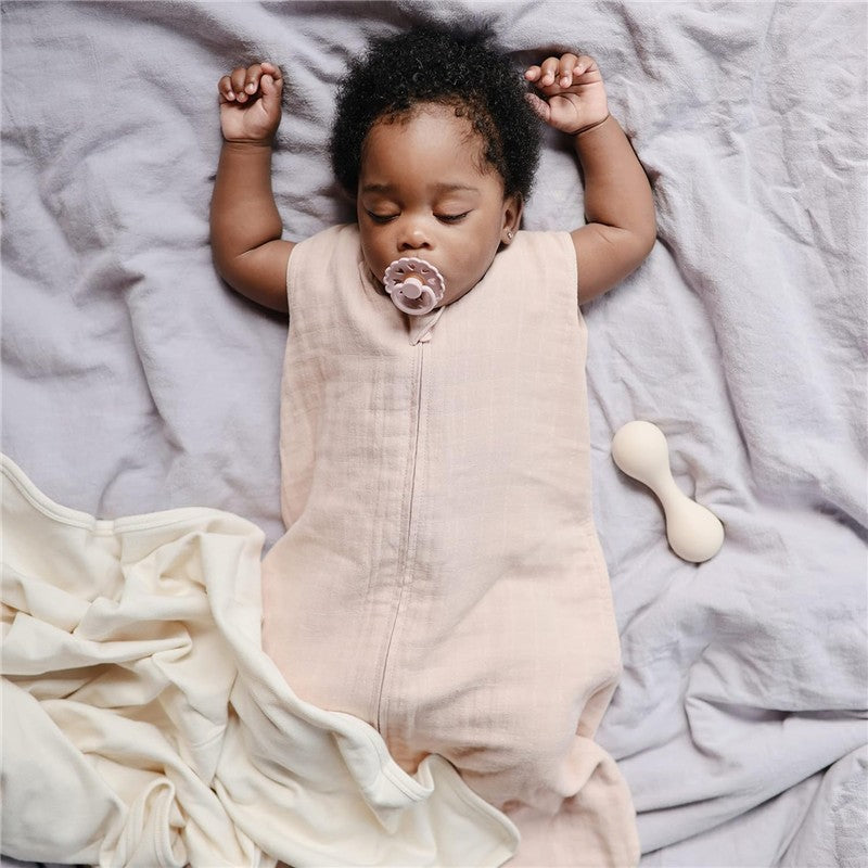 Infant sleeping peacefully in a blush-colored Mushie muslin sleep bag, with a pacifier, surrounded by soft fabrics.