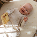 Smiling baby in a blush Mushie Muslin Sleep Bag, holding a yellow toy on a patterned sleep surface.
