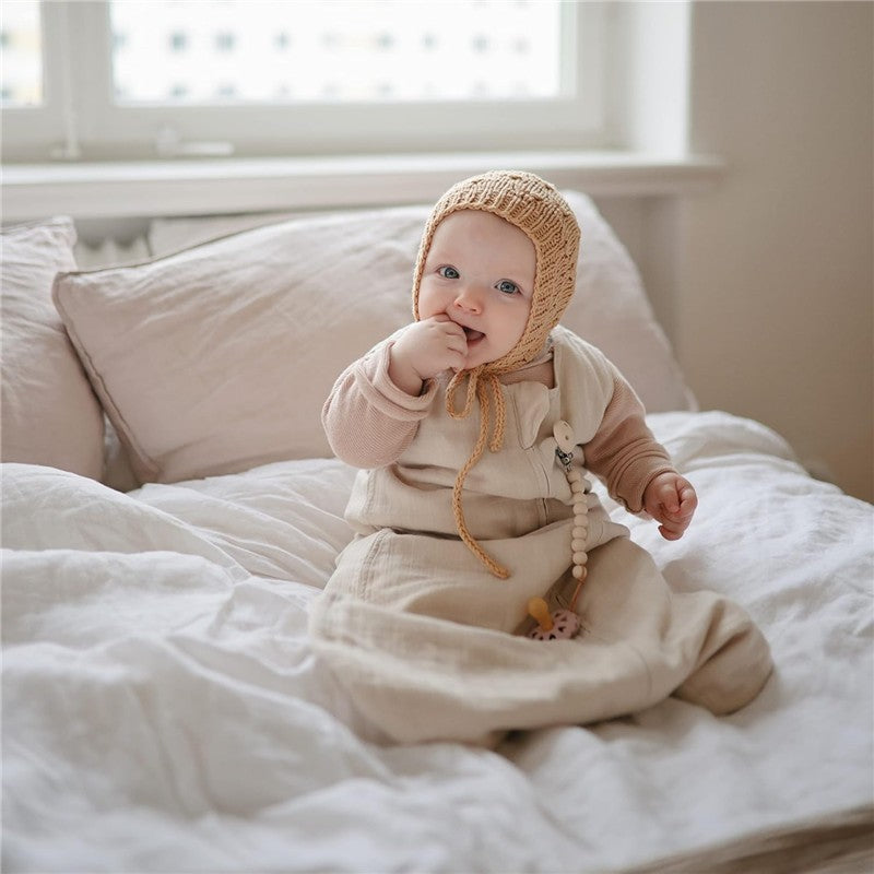 Baby wearing a beige Mushie Muslin Sleep Bag in fog color, sitting on a bed, smiling and sucking on their fingers.