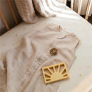 Mushie Muslin Sleep Bag in Fog color, a tan soft fabric sleep sack, alongside a brown pacifier and a yellow silicone toy on a textured crib mattress.