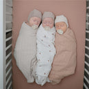 Three newborns swaddled in different Mushie muslin blankets, resting on a blush-colored crib mattress.