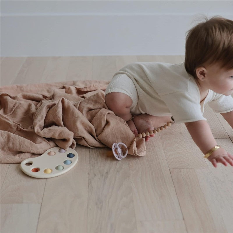 Mushie Muslin Swaddle Blanket in blush color, with a baby crawling on soft wooden floor, surrounded by toys.