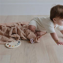 Mushie Muslin Swaddle Blanket in blush color, with a baby crawling on soft wooden floor, surrounded by toys.