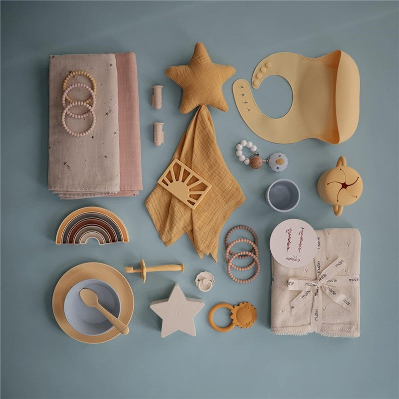 Mushie Muslin Swaddle Blanket in blush with various baby accessories like bibs, teething rings, plates, and toys arranged on a light blue background.