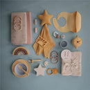 Mushie Muslin Swaddle Blanket in blush with various baby accessories like bibs, teething rings, plates, and toys arranged on a light blue background.
