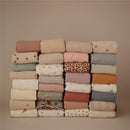 Mushie Muslin Swaddle Blankets in various colors and patterns neatly stacked, highlighting the blush variant.