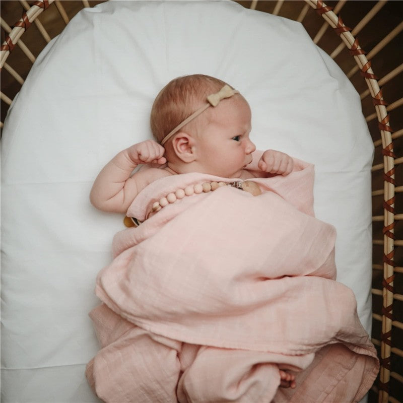 Infant swaddled in a pink muslin blanket with a wooden bead necklace, resting in a wicker bassinet.