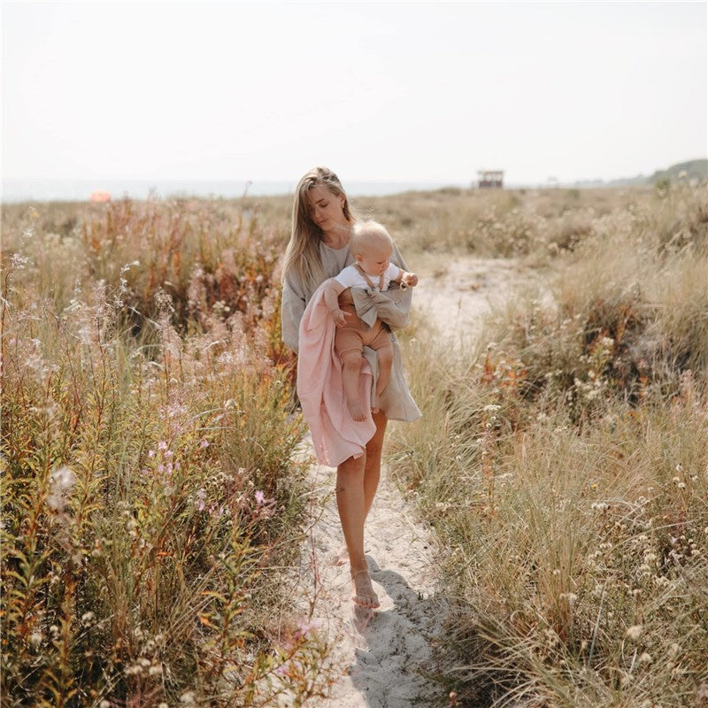 A woman walks along a sandy path carrying a baby wrapped in a pink swaddle blanket amidst tall grass and colorful wildflowers.