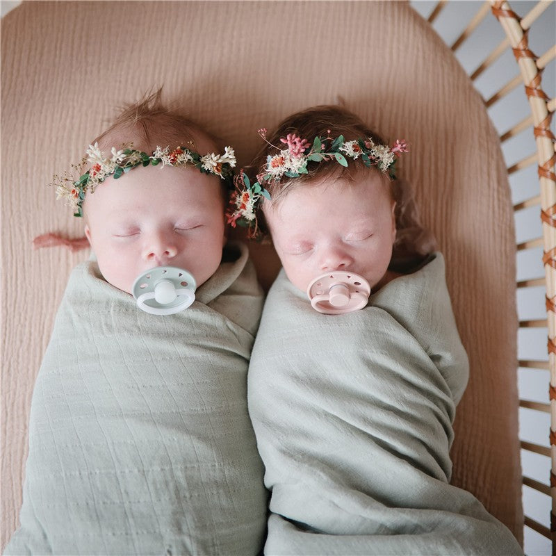 Twin babies sleeping peacefully, swaddled in sage green Muslin blankets, with floral headbands and pacifiers.