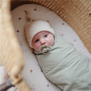 Baby wrapped in a sage green Mushie organic cotton muslin swaddle blanket, wearing a light beanie and pacifier, in a woven bassinet.
