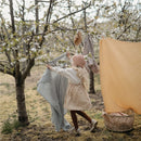Child in a pink hat hanging a sage-colored organic cotton muslin swaddle on a tree branch, surrounded by blooming flowers.