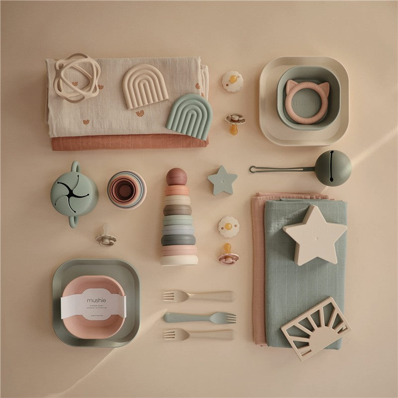 Mushie Organic Cotton Muslin Swaddle Blanket in sage, surrounded by various feeding accessories and toys on a neutral backdrop.