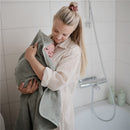 Mother holds a wrapped baby in a moss green hooded towel, in a cozy bathroom setting.