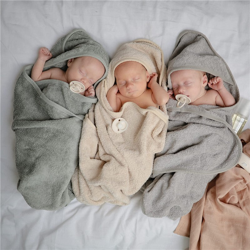 Three wrapped infants asleep in soft hooded towels in moss green, beige, and gray colors, each with a pacifier.
