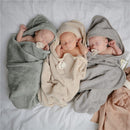 Three wrapped infants asleep in soft hooded towels in moss green, beige, and gray colors, each with a pacifier.