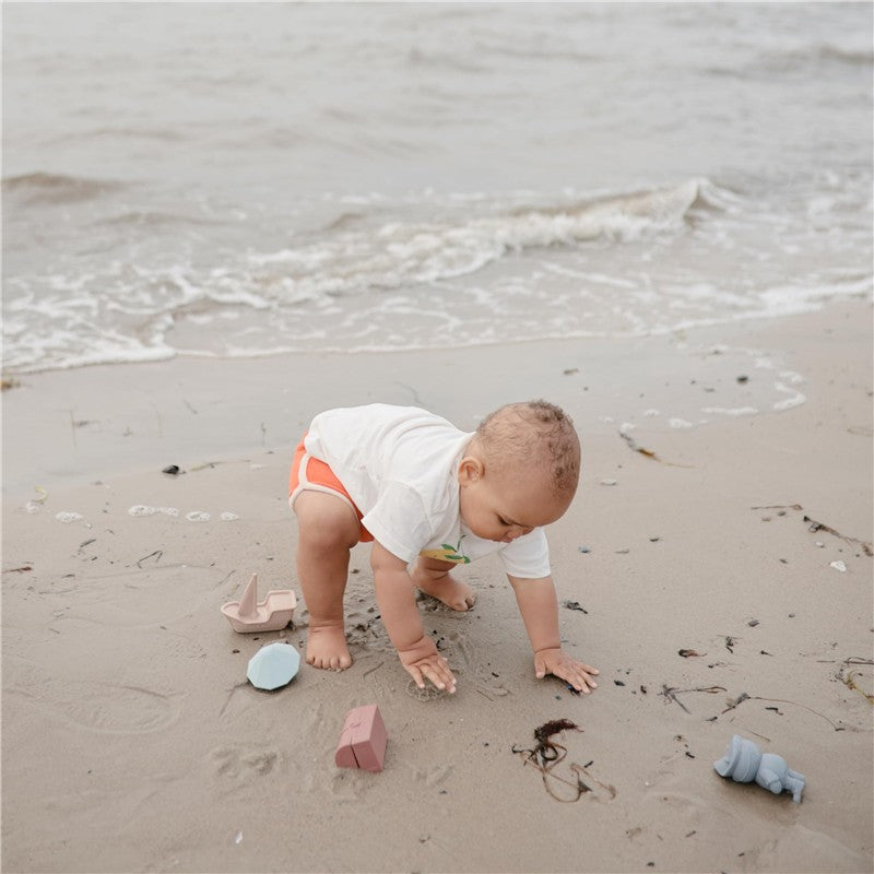 Toddler playing on the beach with a Mushie Pirate Mold Free Bath Play Set, featuring colorful molds scattered on the sand.