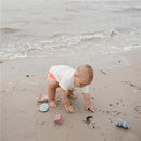 Toddler playing on the beach with a Mushie Pirate Mold Free Bath Play Set, featuring colorful molds scattered on the sand.