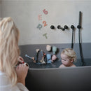 Mushie Silicone Pour and Play Bath Set in use, featuring a child playing in a grey bathtub with colorful silicone cups and a wall decorated with letters.