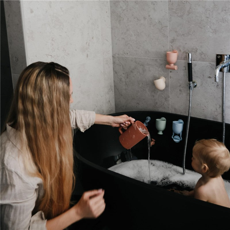 A woman pours water from a clay-colored cup into a black bathtub, where a small child sits among bubbles and colorful bath toys.