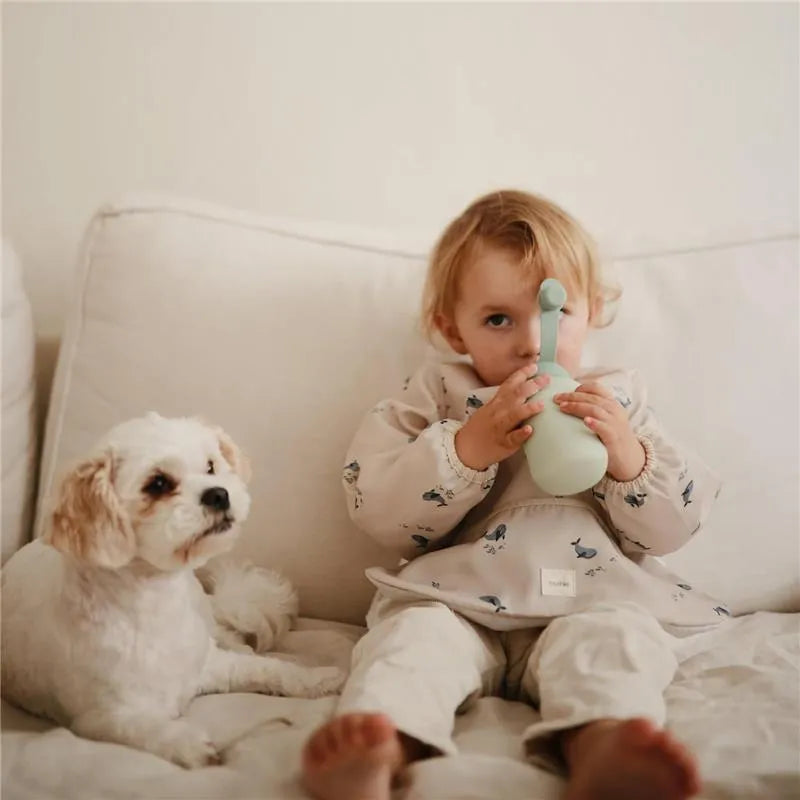 A child sits on a couch, holding a green Mushie Puree Pouch, with a small white dog beside them.