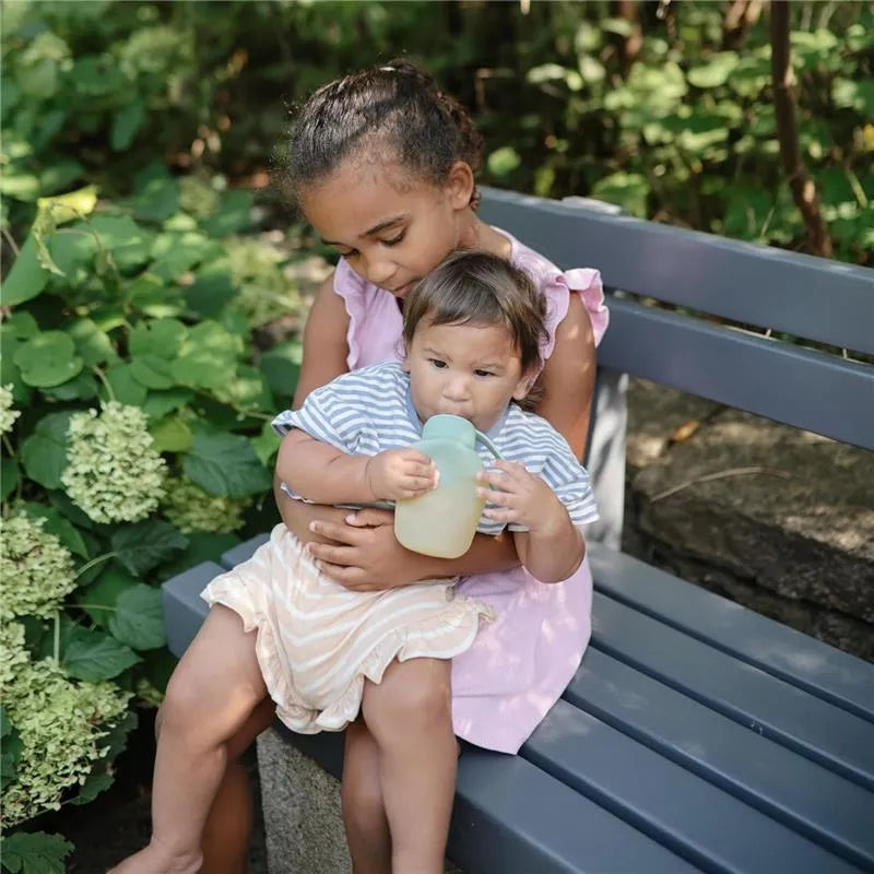 A child in a pink dress sits on a bench, holding a baby in striped clothing who is drinking from a Mushie puree pouch.