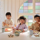 Three children enjoy snacks on a table featuring the Mushie Silicone Round Plate Dinnerware Set in smoke color, beside colorful cups.