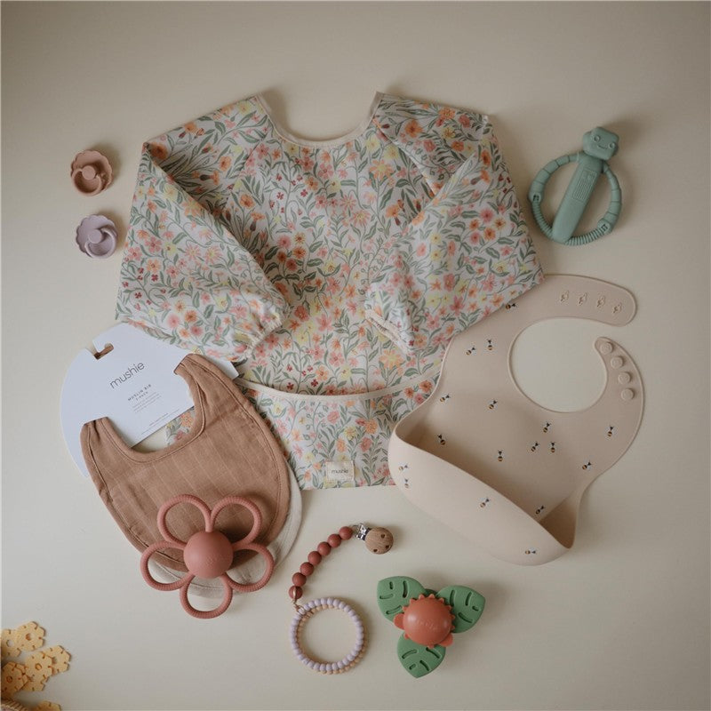 Mushie silicone baby bib in beige with bee designs, alongside a floral long-sleeve top and various teething toys.