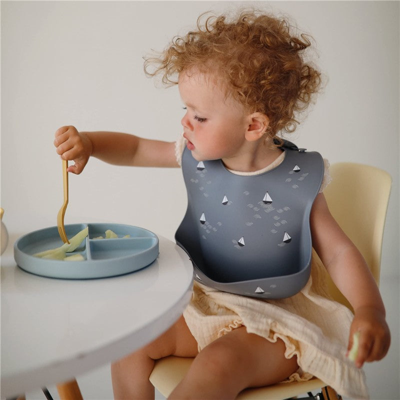 Child wearing a gray silicone baby bib with boat designs, using a fork to eat from a divided blue plate.