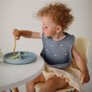 Child wearing a gray silicone baby bib with boat designs, using a fork to eat from a divided blue plate.