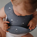 Silicone baby bib featuring a boat design, being worn by an infant exploring its pocket with small hands.