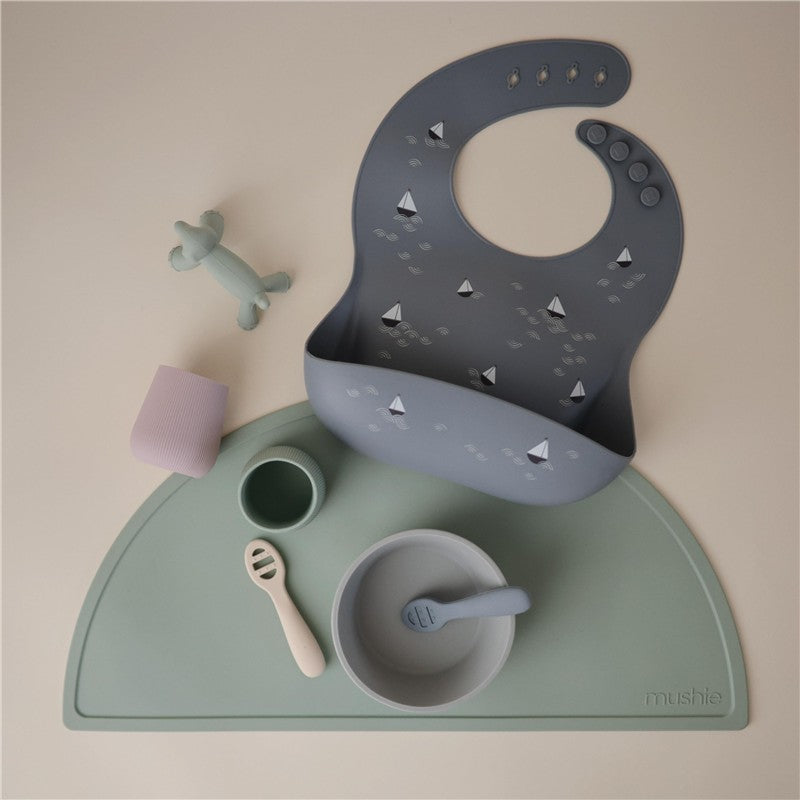 Silicone baby bib in gray with boat designs, alongside various feeding items on a green placemat.