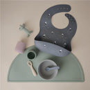 Silicone baby bib in gray with boat designs, alongside various feeding items on a green placemat.