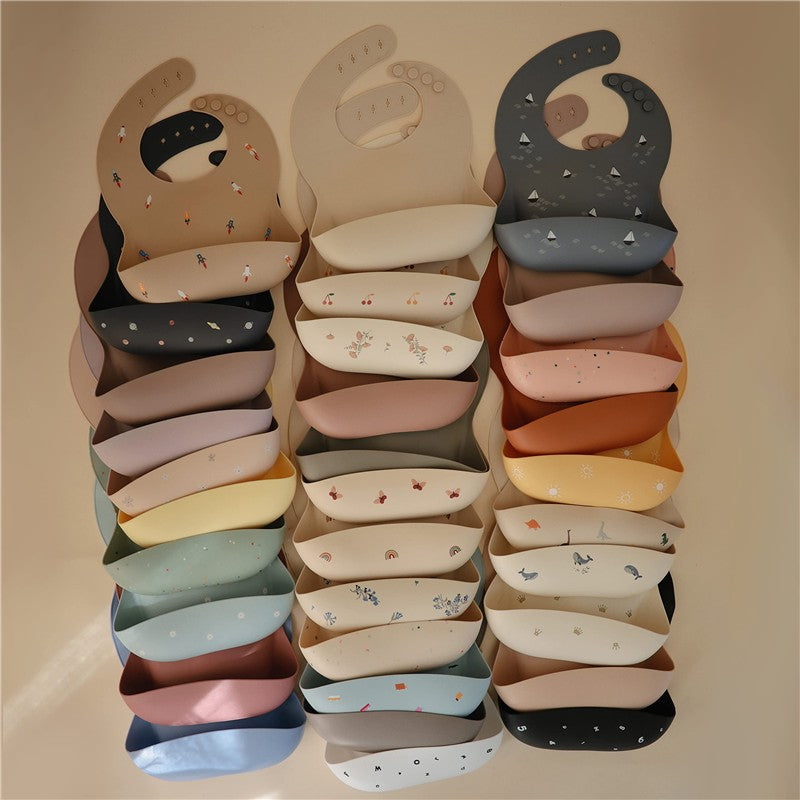 Silicone baby bibs in various colors and prints, including designs with boats and whimsical patterns, stacked together.