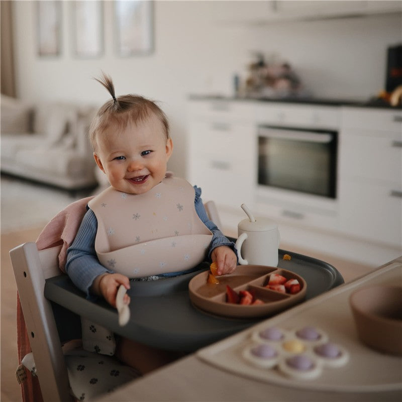 A smiling baby wearing a Mushie Silicone Baby Bib in Daisy, sitting at a high chair with fruit on a plate.