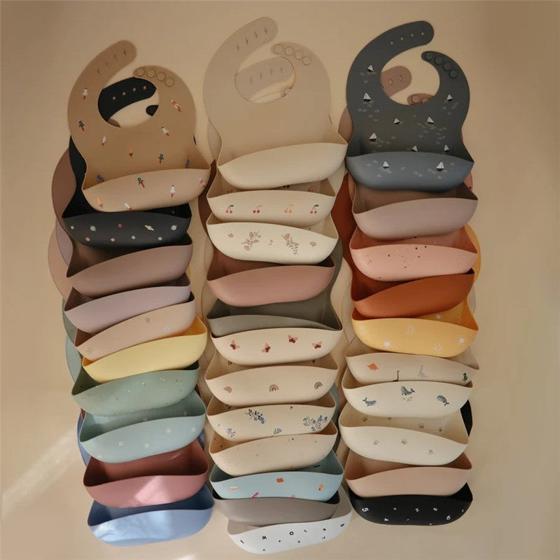 Mushie Silicone Baby Bibs in multiple colors and patterns, featuring playful designs like stars and flowers, stacked neatly.