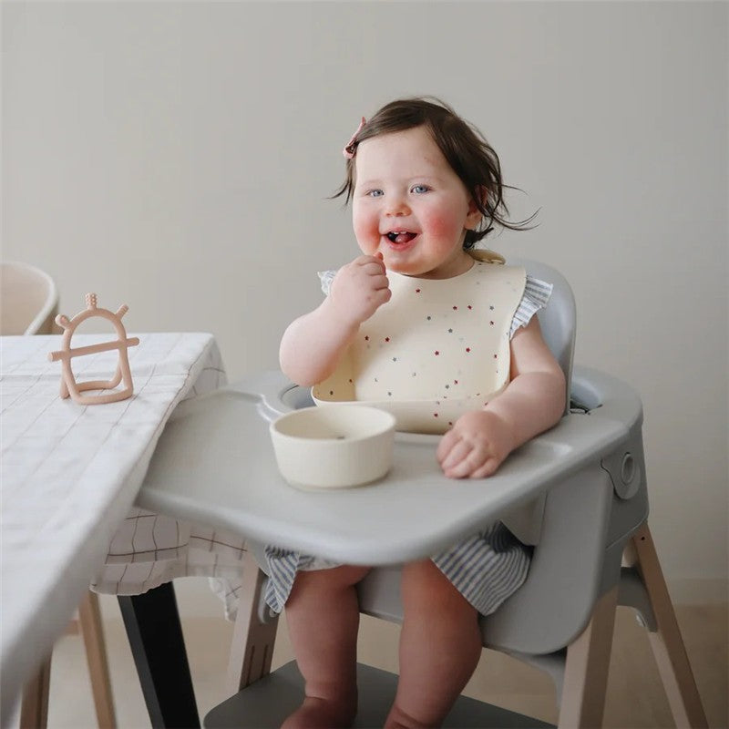 A smiling baby wearing a Mushie silicone bib with stars sits in a high chair, eating from a bowl on the tray.