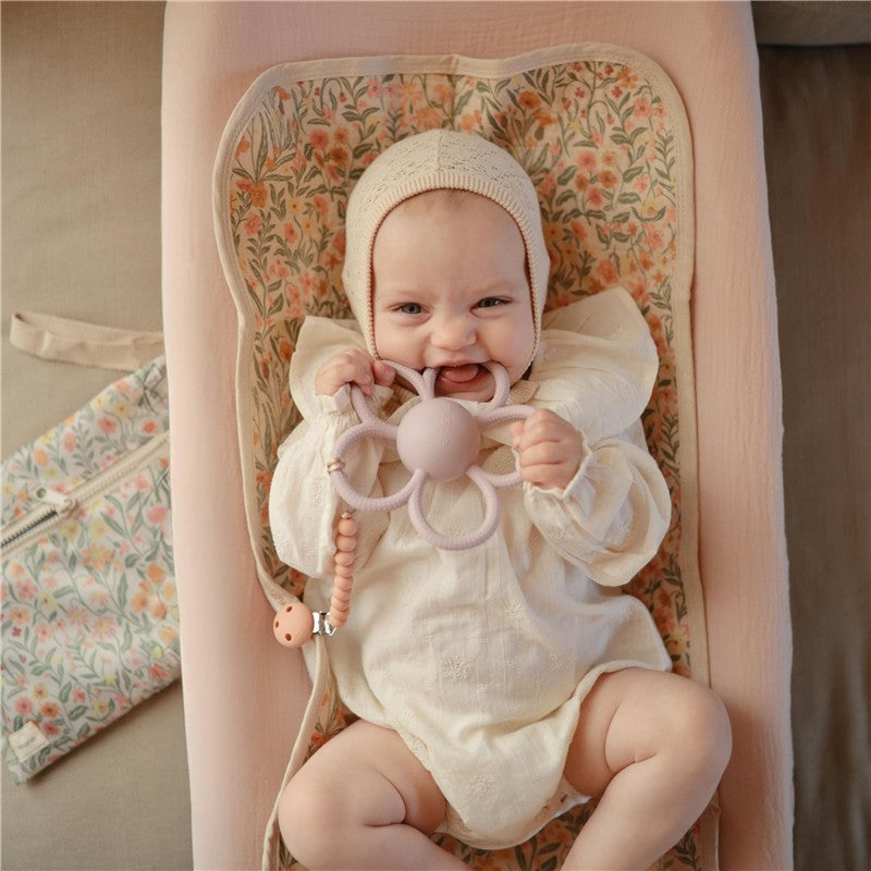 Baby smiling while holding a soft lilac Mushie Daisy Rattle Teether, seated on a floral-patterned infant seat.
