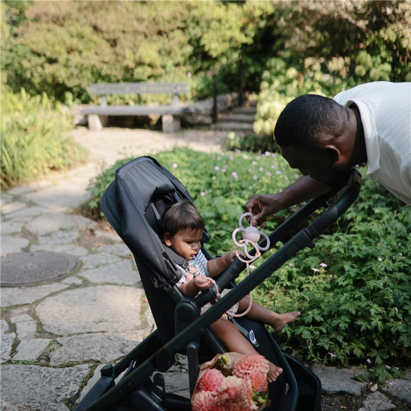 A child in a stroller holds a Mushie Daisy Rattle Teether in soft lilac while an adult engages with them in a garden setting.