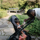 A child in a stroller holds a Mushie Daisy Rattle Teether in soft lilac while an adult engages with them in a garden setting.