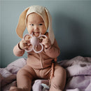 Baby wearing a pink outfit and bunny-ear hat, holding a soft lilac Mushie Daisy rattle teether while sitting on a purple blanket.