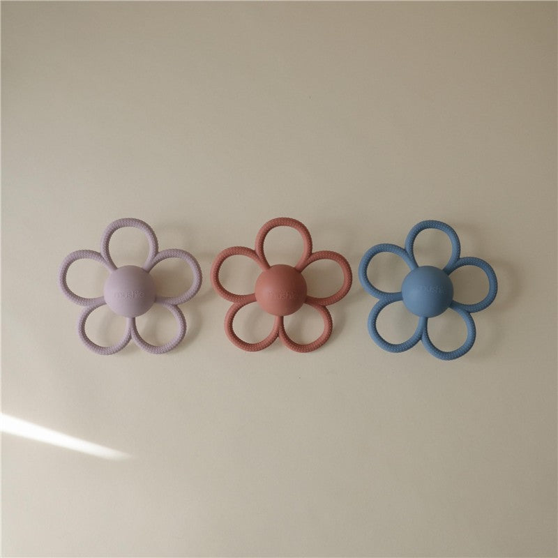 Mushie Daisy Rattle Teether in soft lilac, alongside petal-shaped designs in pink and blue, lying on a light background.