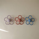 Mushie Daisy Rattle Teether in soft lilac, alongside petal-shaped designs in pink and blue, lying on a light background.