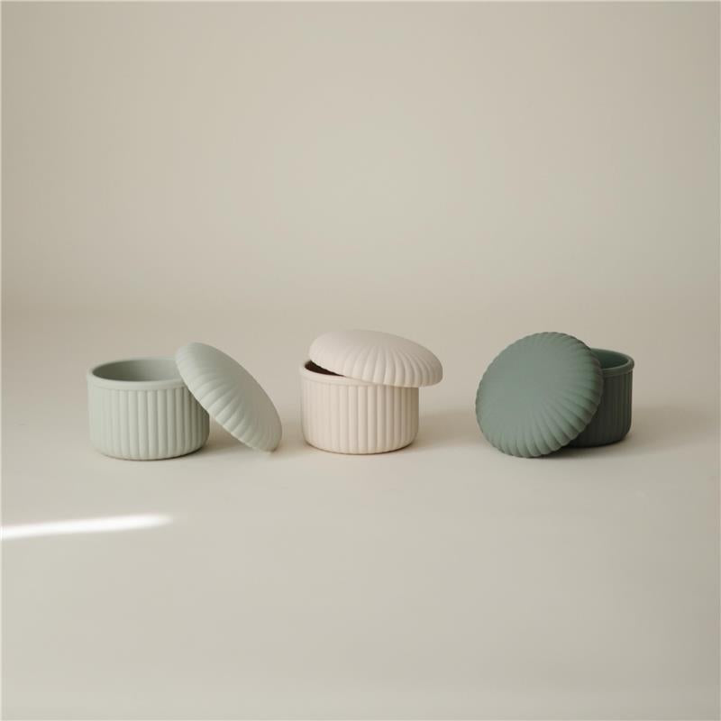 Mushie silicone baby food containers in green and cream, featuring ribbed designs and matching lids.