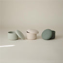 Mushie silicone baby food containers in green and cream, featuring ribbed designs and matching lids.